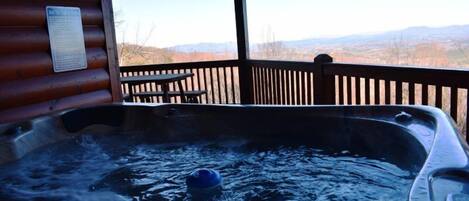 A hot tub with a wooden deck and a table, overlooking a mountainous landscape.