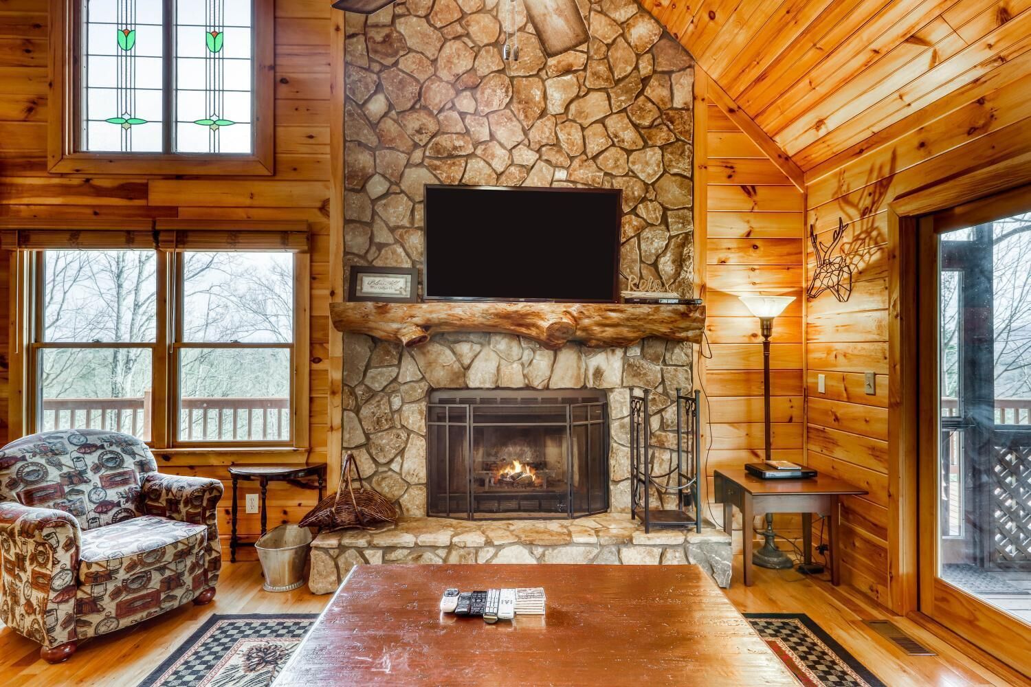 A cozy cabin interior with a stone fireplace, wooden walls, and a large window.