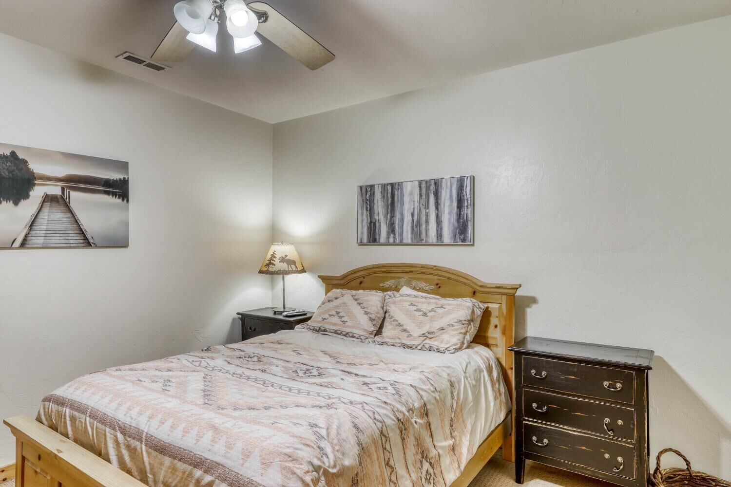 A bedroom with a wooden bed, a nightstand with a lamp, a dresser, and two wall art pieces.