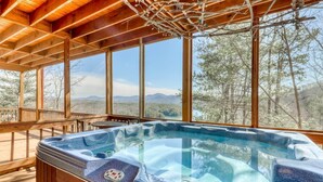 Vasca idromassaggio su un pontile di legno con ampie finestre che si affacciano su una foresta e montagne.