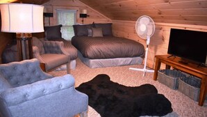 Quarto de sótão acolhedor com uma cama, duas poltronas, um abajur, um ventilador, uma televisão e um tapete preto grande.