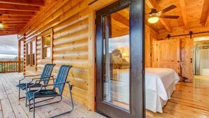 Una cabaña de madera con terraza, dos sillas, una cama y un ventanal grande.