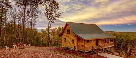 A wooden cabin with a porch, surrounded by trees and a leaf-covered ground.