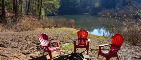 Three red Adirondack chairs arranged around a campfire in a natural setting with a lake and forest in the background.
