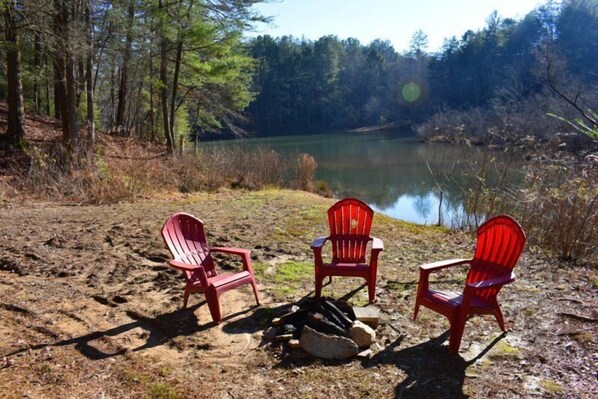 Three red Adirondack chairs arranged around a campfire in a natural setting with a lake and forest in the background. - Misty Lake Lodge (Blue Ridge)