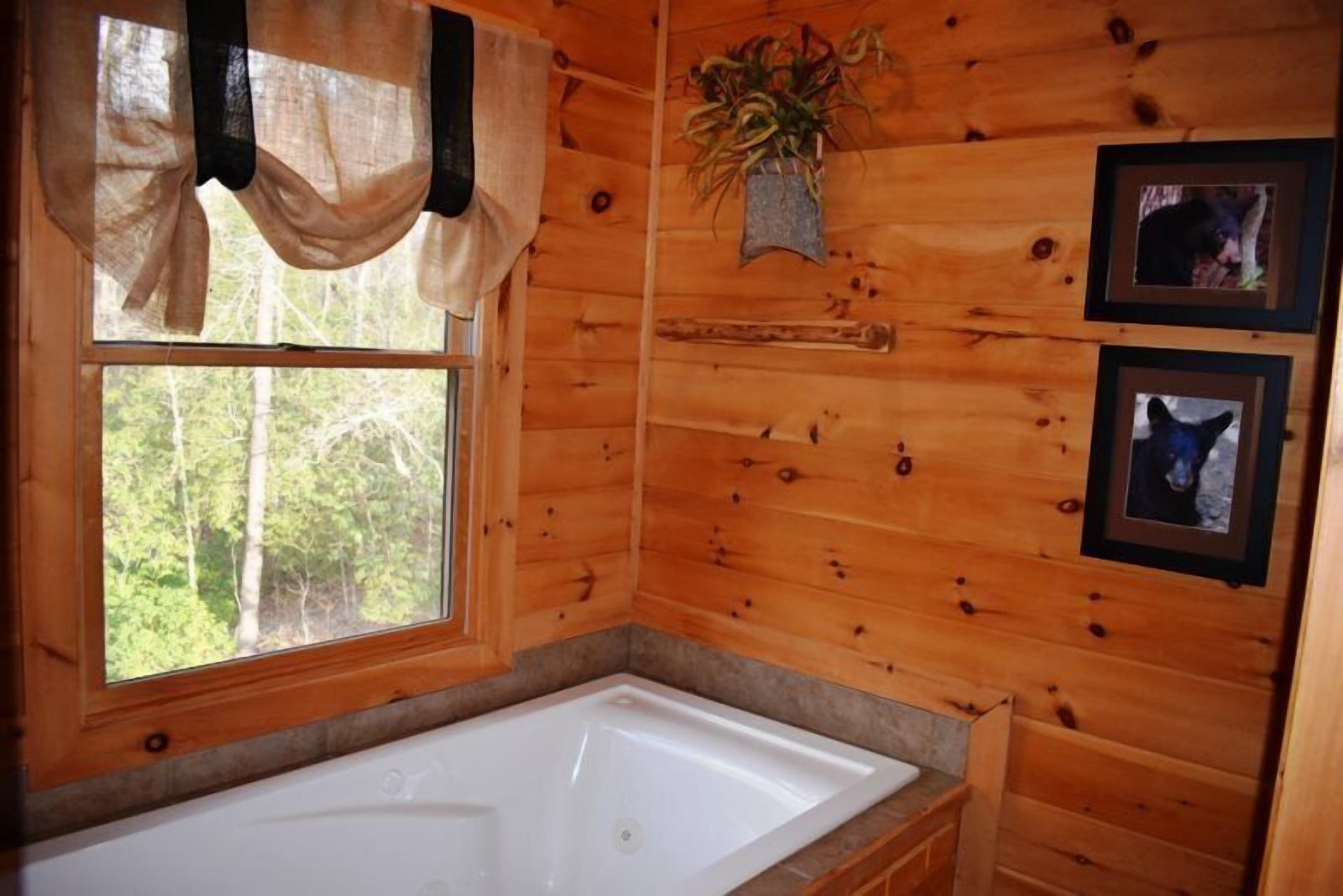 Casa de banho de uma cabana de madeira com uma banheira grande, uma janela com cortinas leves e quadros nas paredes.