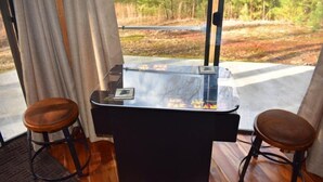 Une pièce avec une table à plateau de verre, deux tabourets en bois et une vue sur une forêt, visible par une fenêtre.