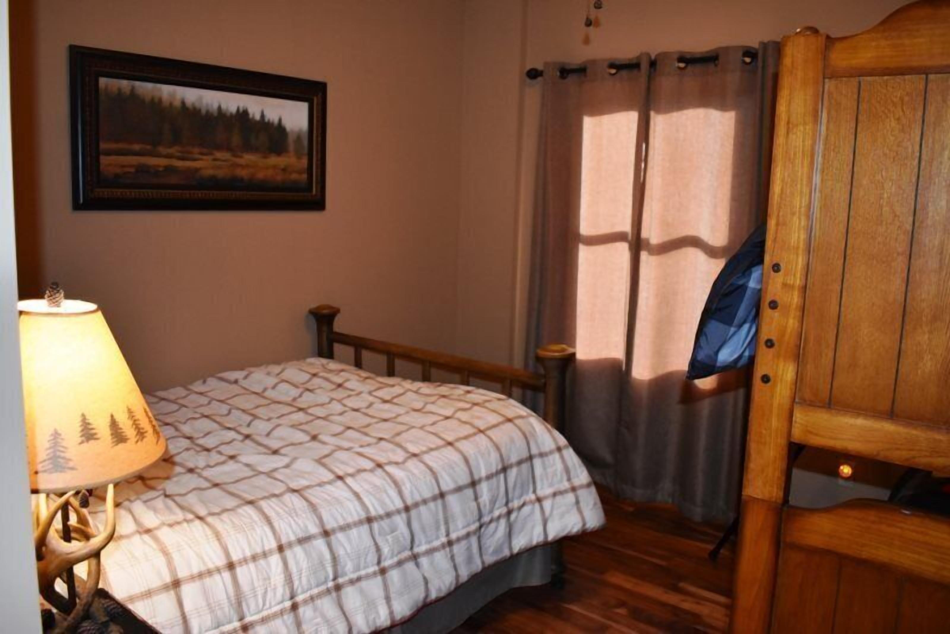 A bedroom with a bed, a nightstand, a lamp, a wooden chair, a window with curtains, and a framed picture on the wall.