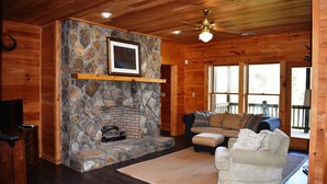 A cozy cabin interior with a stone fireplace, wooden walls, a ceiling fan, and a comfortable seating area.