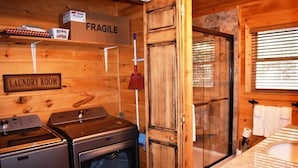 Un cuarto de lavandería con lavadora y secadora, una caja delicada sobre una estantería y una zona de ducha con puerta de vidrio.