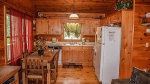 Eine Holzhütte mit Küchenbereich, ausgestattet mit einem Holztisch, Stühlen, einem Kühlschrank und einem Fenster mit Vorhängen.