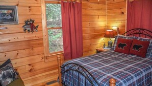Ein Schlafzimmer einer Holzhütte mit einer Karo-Bettdecke, einem Nachttisch mit Lampe und einem Fenster mit roten Vorhängen.