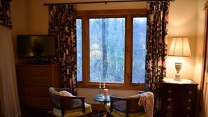 Une pièce chaleureuse avec un téléviseur posé sur un meuble en bois, deux fauteuils avec des coussins, une petite table avec une bougie et une lampe sur une table d’appoint.