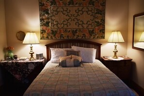A neatly made bed with a floral-patterned bedspread, flanked by two bedside tables with lamps, a large tapestry on the wall, and a mirror on the right.