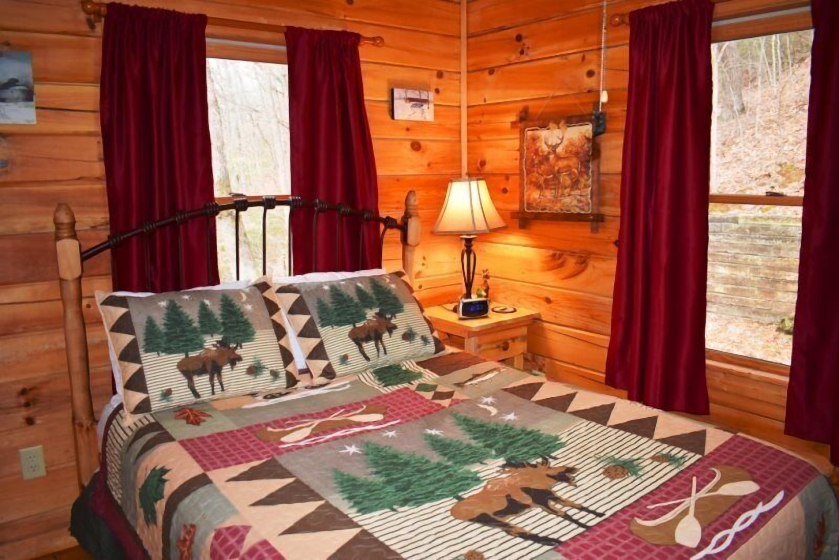 A log cabin bedroom with a bed featuring a moose-themed quilt, red curtains, and a nightstand with a lamp.