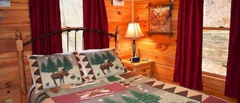A log cabin bedroom with a bed featuring a moose-themed quilt, red curtains, and a nightstand with a lamp.