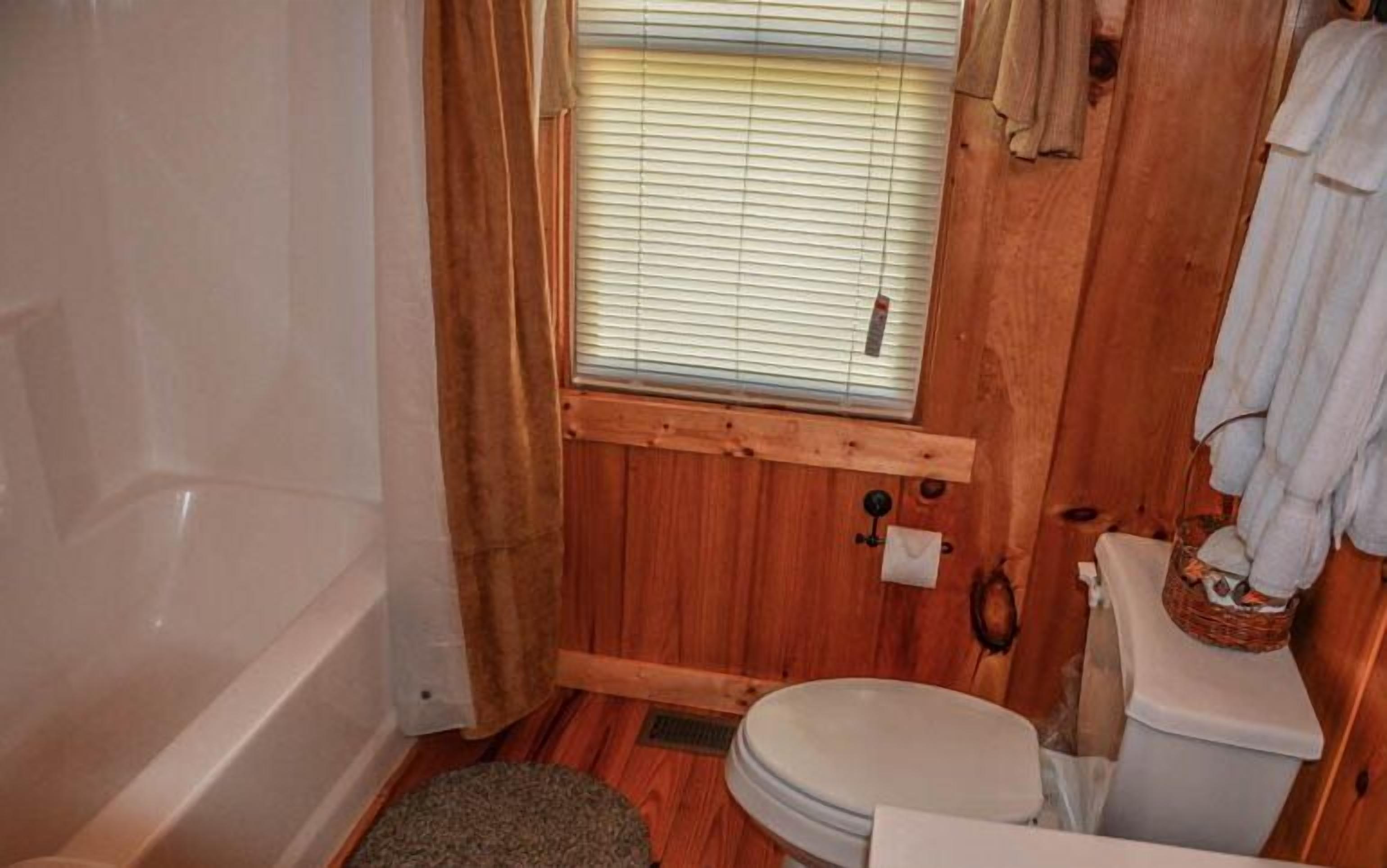 A compact bathroom with a wooden interior, a bathtub, a toilet, and a window with blinds.