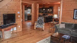 A cozy cabin interior with a wooden floor, a television, a sofa, a patterned armchair, and a kitchen area in the background. - Run About Trout Lodge (Blue Ridge)