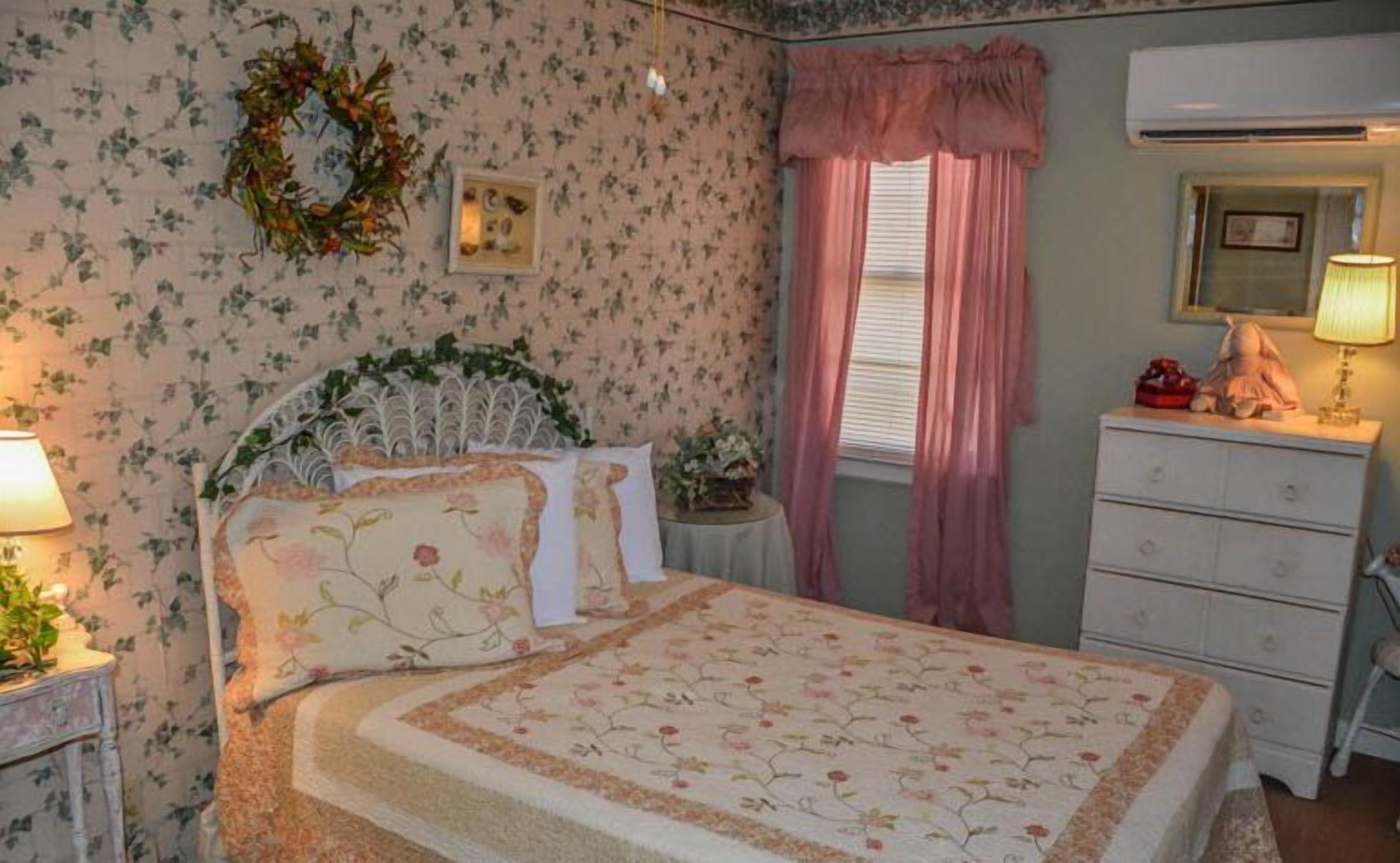 Un dormitorio con papel tapiz floral, una cama con ropa de cama floral, una mesita de noche con lámpara, un aparador y una ventana con cortinas rosas.