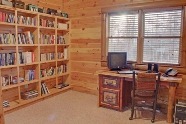 A room with a wooden bookshelf filled with books, a desk with a computer, and a window with blinds. - Safe Haven (Morganton)