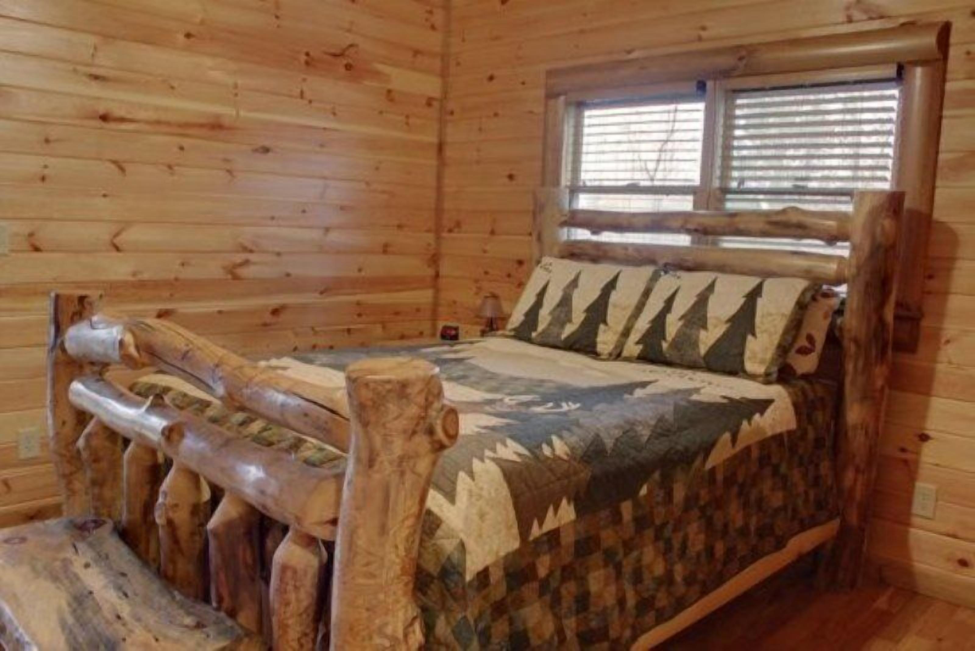 Interno di una baita in legno con un letto, una coperta a tema bosco, una finestra con persiane e una testiera in legno rustico.