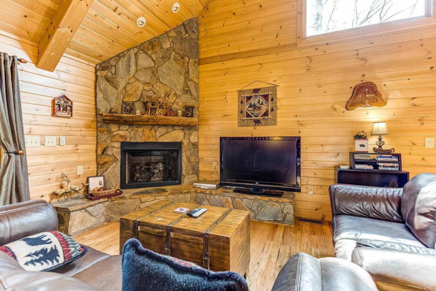 L’intérieur chaleureux d’un chalet avec une cheminée en pierre, un téléviseur, des meubles en cuir et un plafond en bois.