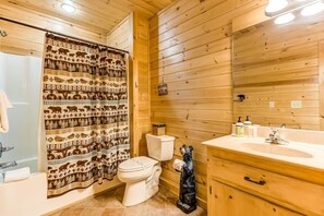 A wooden cabin bathroom with a patterned shower curtain, a toilet, a sink with a mirror, and a small statue. - Mountain Tops Serenity (Blue Ridge)