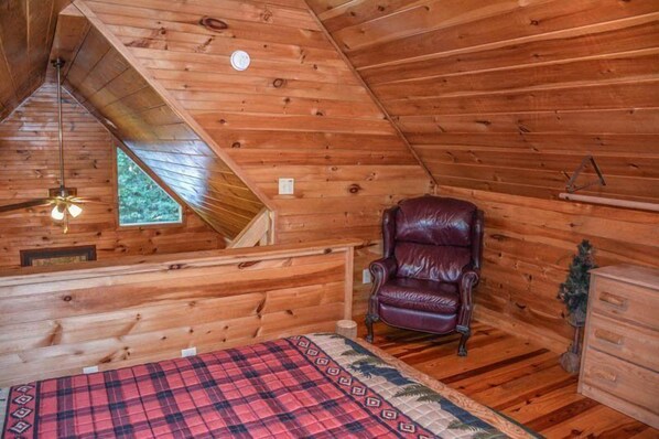 A wooden cabin room with a sloped ceiling, a leather chair, a bed with a plaid blanket, and a small window. - Hackers Hideaway (Blue Ridge)