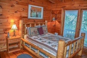 A wooden cabin bedroom with a bed, a nightstand, a lamp, and a window with blinds. - Cherry Acre (Blue Ridge)