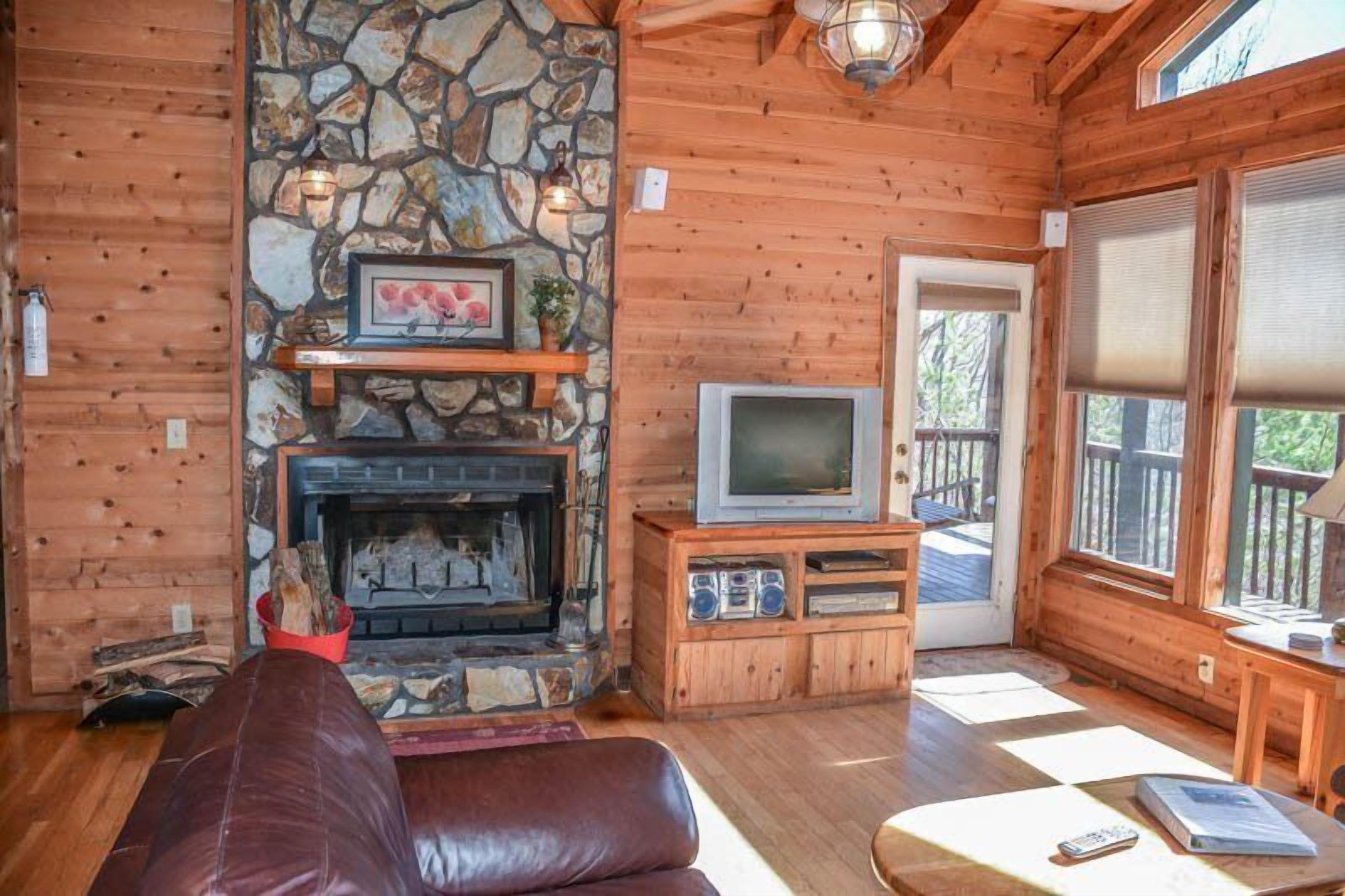 L’intérieur chaleureux d’un chalet avec une cheminée en pierre, des murs en bois, un canapé en cuir et un téléviseur posé sur un meuble en bois.