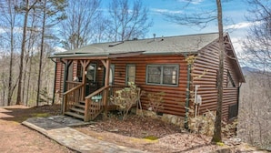 Une cabane rustique en rondins de bois, dotée d’un porche, d’un chemin en pierre et offrant une vue sur la forêt environnante.