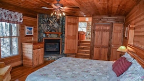 Une chambre confortable, aménagée dans le style d’un chalet, avec un grand lit, des murs en bois, une cheminée en pierre et un ventilateur de plafond.