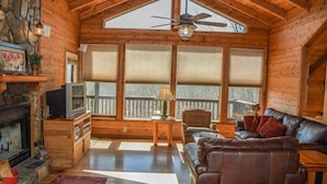 L’intérieur d’un chalet confortable, avec une cheminée, un téléviseur, un canapé et de grandes fenêtres.