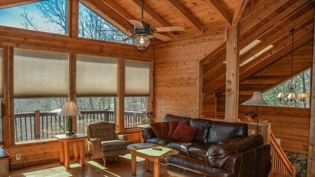 L’intérieur d’un chalet accueillant, avec un plancher en bois, un canapé en cuir, une table à manger et de grandes fenêtres équipées de stores.