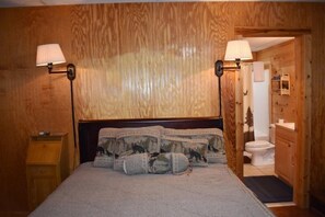 A wooden-paneled bedroom with a bed, two bedside lamps, a nightstand, and a wooden door leading to a bathroom. - My Mountain Escape (Morganton)