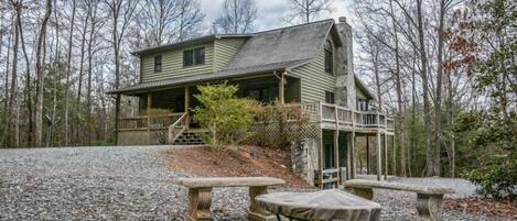 A rustic cabin with a porch, surrounded by trees and gravel.