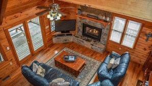 A log cabin interior with a stone fireplace, leather sofas, a television, and a ceiling fan. - My Mountain Escape (Morganton)