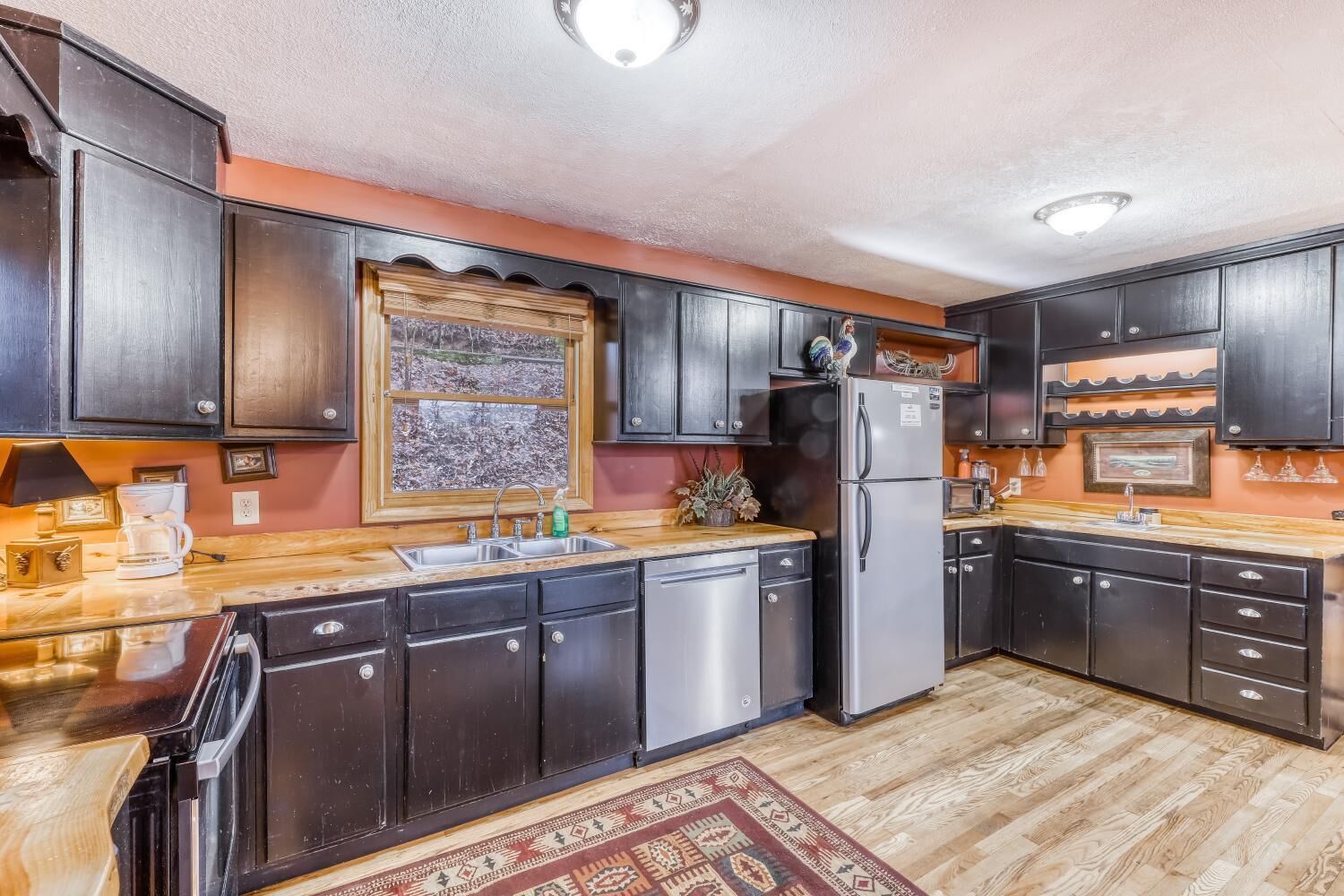 A kitchen with dark wooden cabinets, a stainless steel refrigerator, a wooden countertop, and a window with a view of trees.