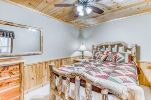 A rustic bedroom with a wooden bed frame, a ceiling fan, a mirror, and a nightstand with a lamp. - Deepwater Lodge (Blue Ridge)