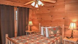 A wooden cabin bedroom with a bed, two bedside lamps, a ceiling fan, and a window with curtains. - Cabin Sweet Cabin (Blue Ridge)