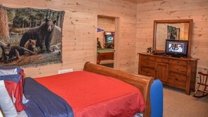 Quarto com teto de madeira, cama com cobertura vermelha, cômoda de madeira, televisão e um mural na parede com tema de urso.