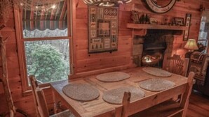 Interior de una cabaña de madera con una mesa de comedor para cuatro, una chimenea y una ventana con vistas a árboles.