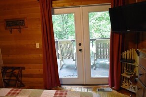 Una habitación con interior de madera, un ventanal corredero que da a un patio exterior con sillas y un televisor a la derecha.