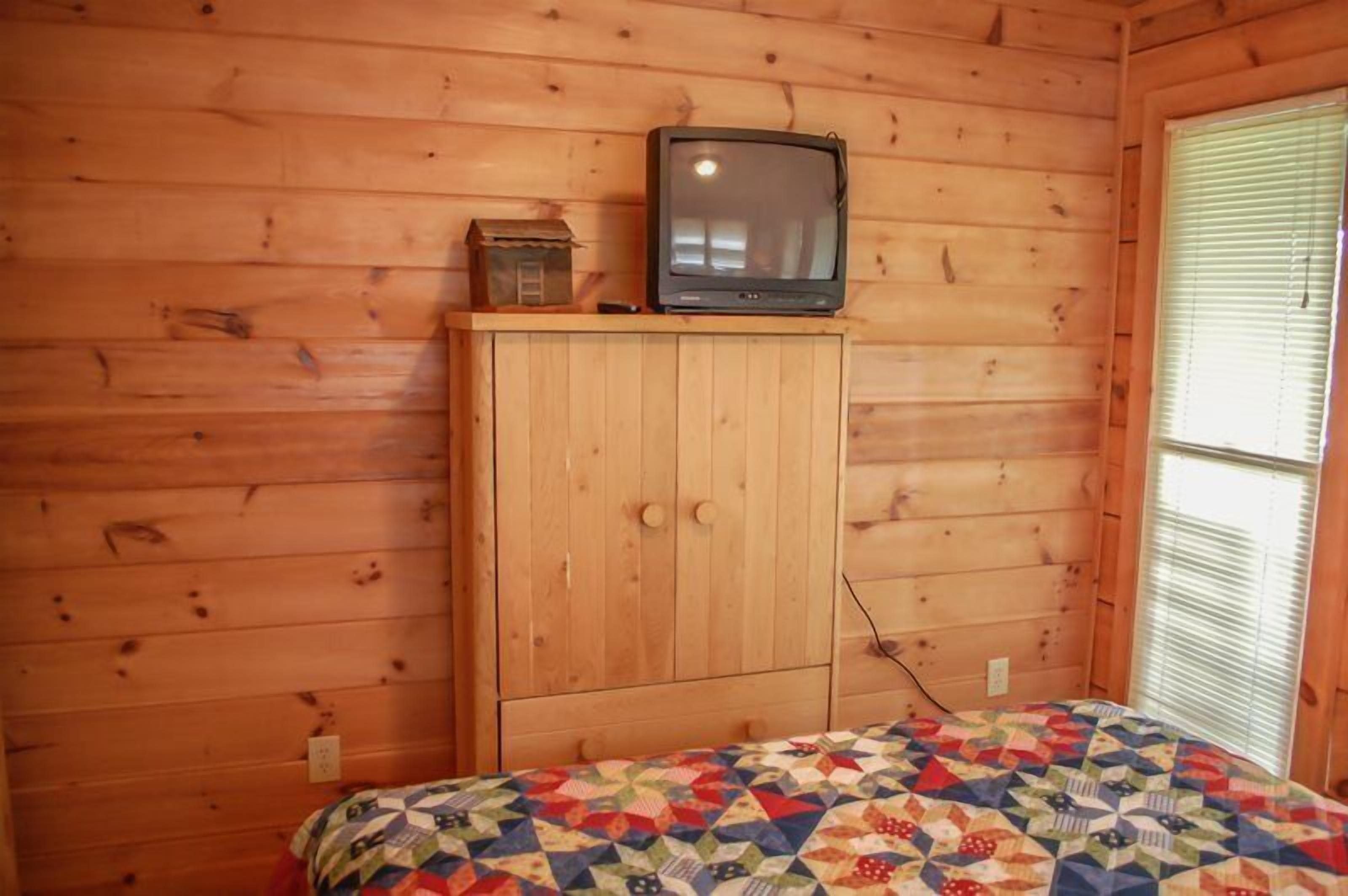 Une pièce de cabane en bois comprenant un lit, un téléviseur posé sur un meuble en bois et une fenêtre avec des stores.