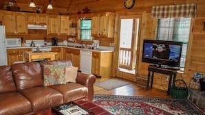 L’intérieur chaleureux d’un chalet avec un canapé en cuir, un pouf rouge, un téléviseur, des meubles en bois, un coin cuisine et un grand tapis.