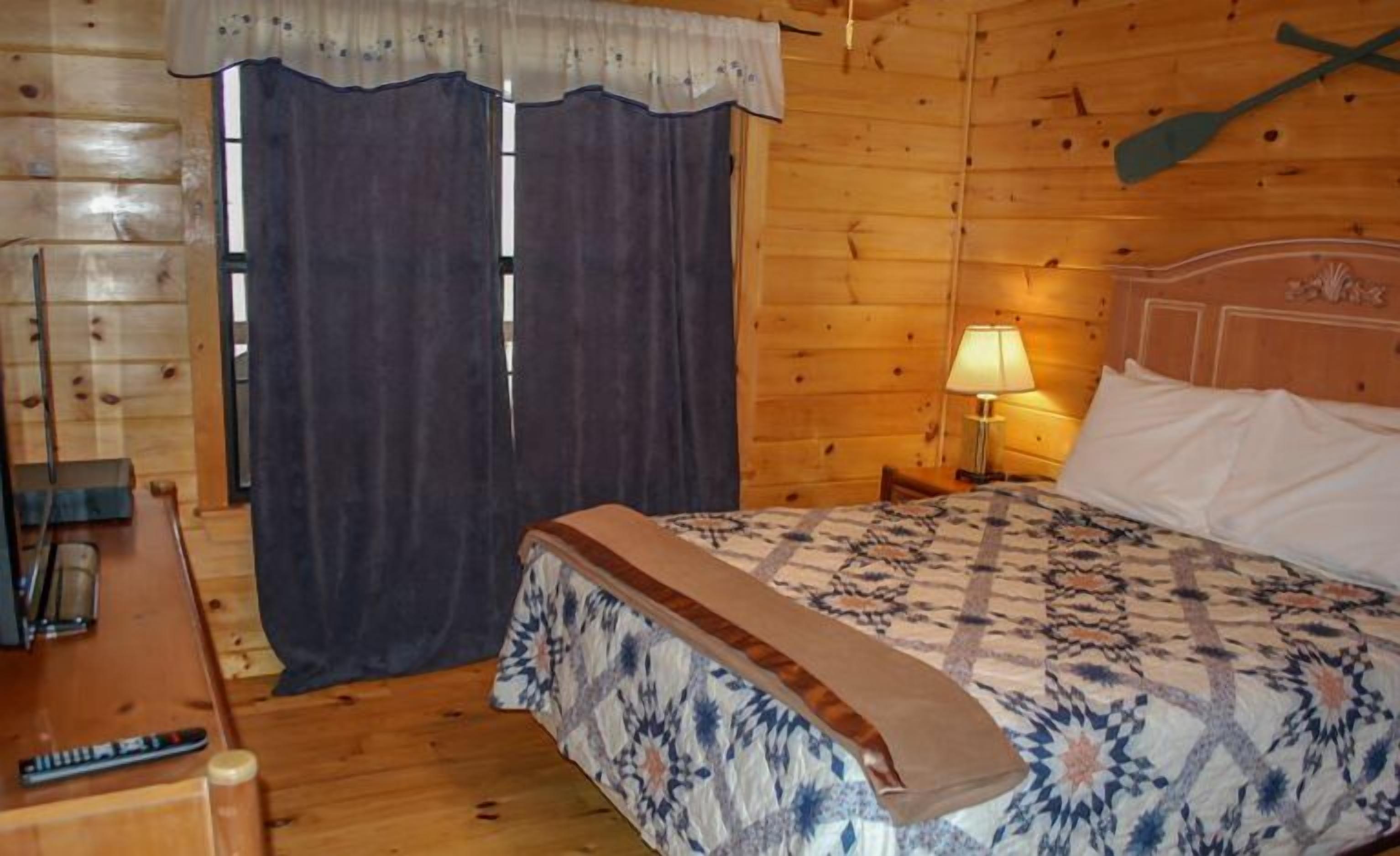 A wooden cabin room with a bed, a nightstand with a lamp, and a window with curtains.