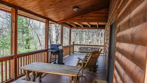 Une terrasse en bois équipée d’un barbecue, d’une table de pique-nique et de chaises, avec vue sur une zone boisée.