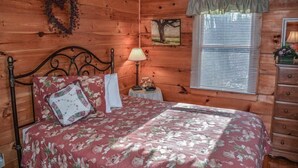 Un dormitorio de cabaña de madera con una cama, una mesita de noche, una lámpara, un aparador, una ventana con persianas y un adorno de pared.