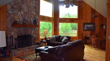 A cozy cabin interior with a stone fireplace, wooden walls, a ceiling fan, and large windows offering a view of the outdoors.
