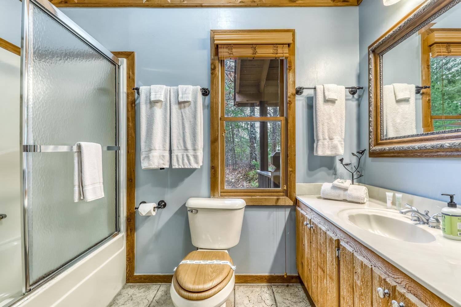 Een badkamer met een douchecabine van glas, een toilet, een wastafel met een opbergmeubel en een raam met uitzicht op bomen.